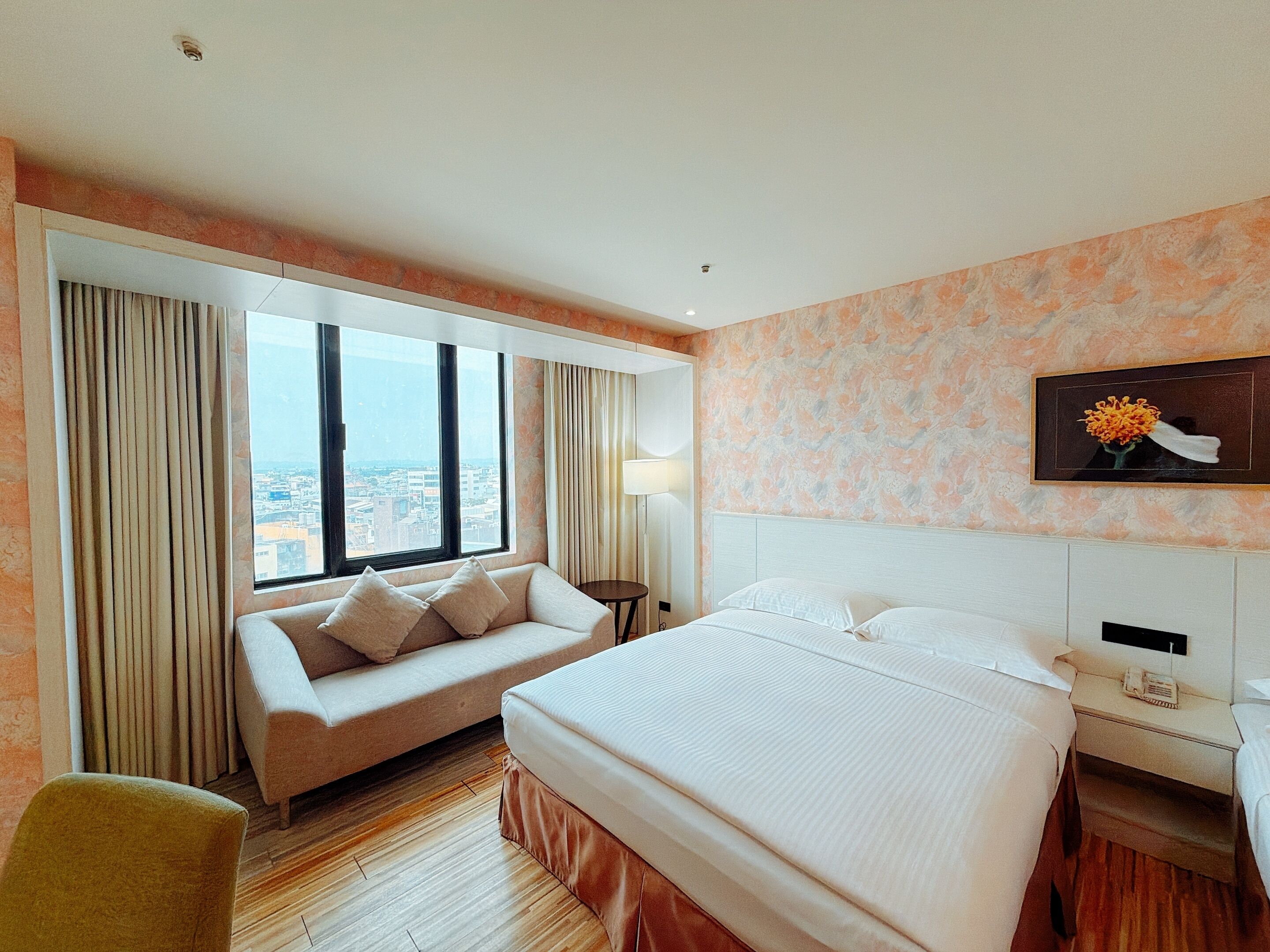 A hotel room with a large bed, a sofa, a small table, a chair, and a large window offering a city view.