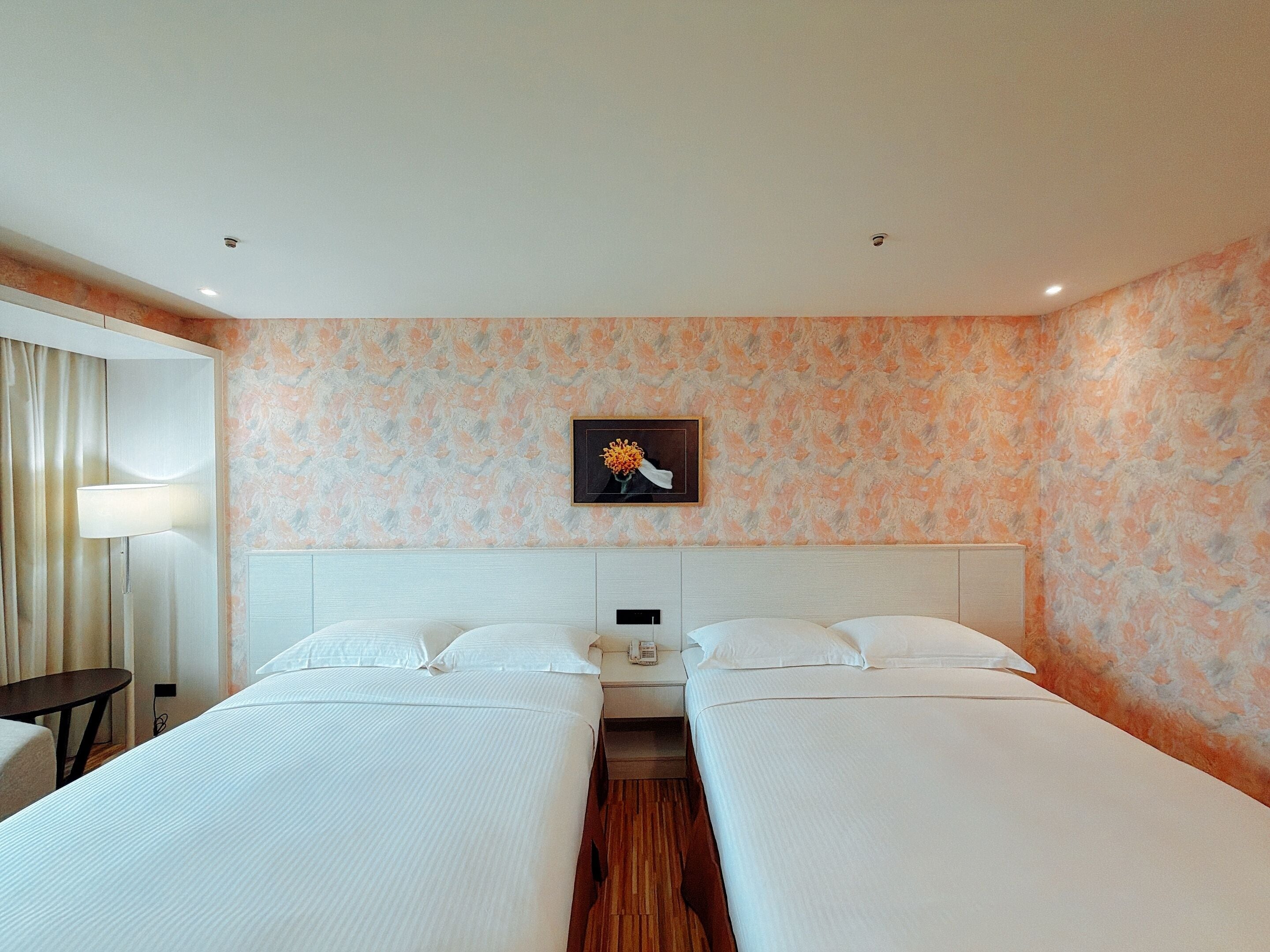 A hotel room with two beds, a bedside table, a lamp, and a floral artwork on the wall.