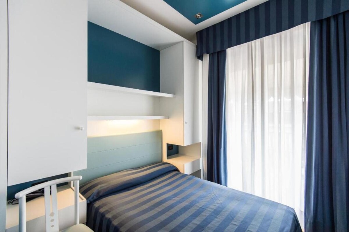 A bedroom with a bed, a bedside table, a nightstand, a wall-mounted shelf, and a window with curtains.