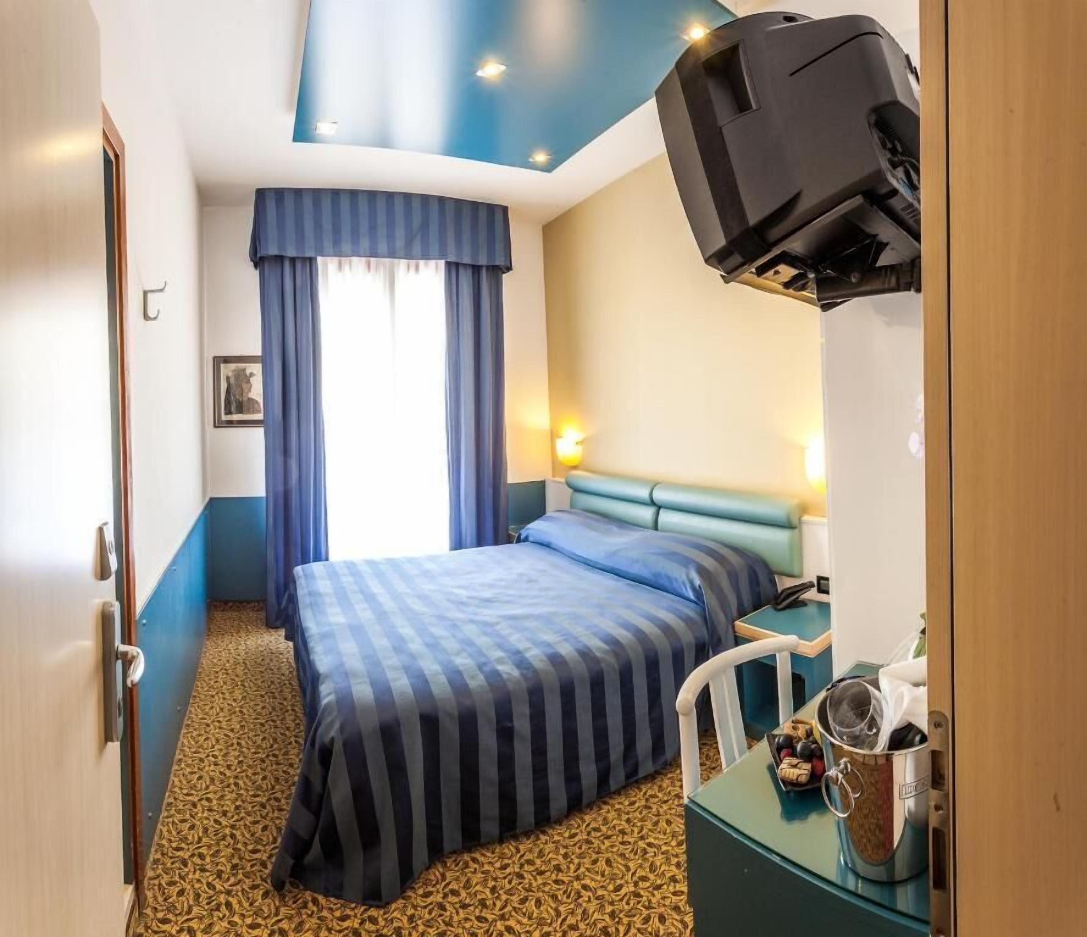 A hotel room with a bed, a desk with a kettle, a television mounted on the wall, and a window with curtains.