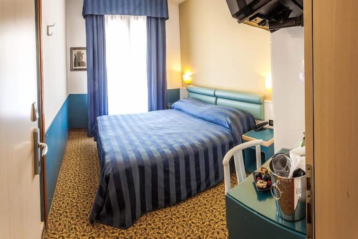 A hotel room with a bed, a desk with a kettle, a television mounted on the wall, and a window with curtains.
