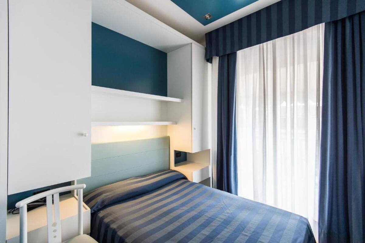 A bedroom with a bed, a bedside table, a nightstand, a wall-mounted shelf, and a window with curtains.