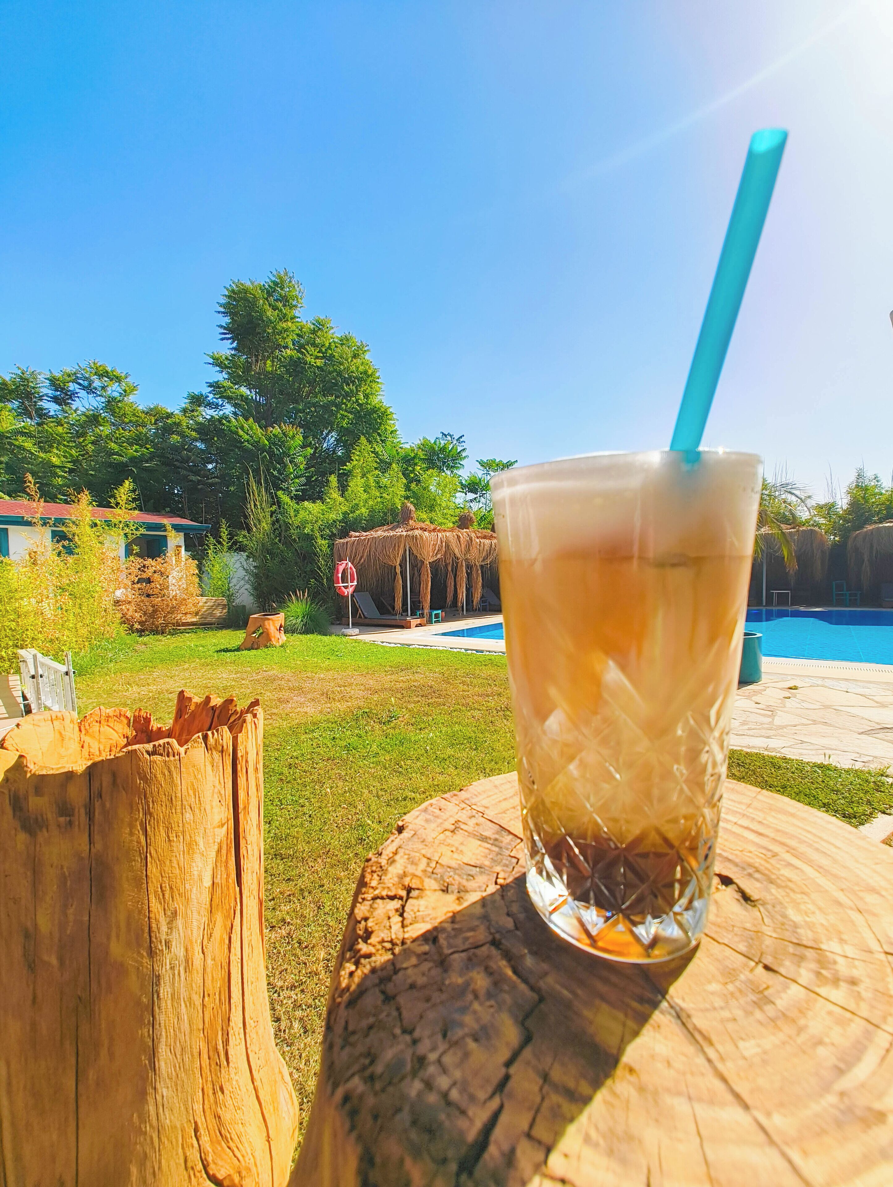 A glass of iced tea on a wooden stump with a blue straw, set against a backyard with a pool and a thatched-roof structure.