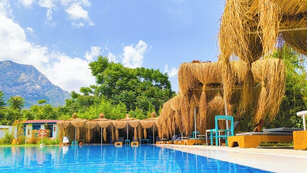A clear blue pool with lounge chairs and thatched umbrellas, surrounded by lush greenery and mountains in the background. - Erendiz Garden Hotel (Kemer)