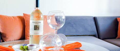 En flaska Angora-vin och ett glas står på ett bord tillsammans med en tallrik med druvor och en liten skål med sås. Allt är placerat på en soffa med orange kuddar.