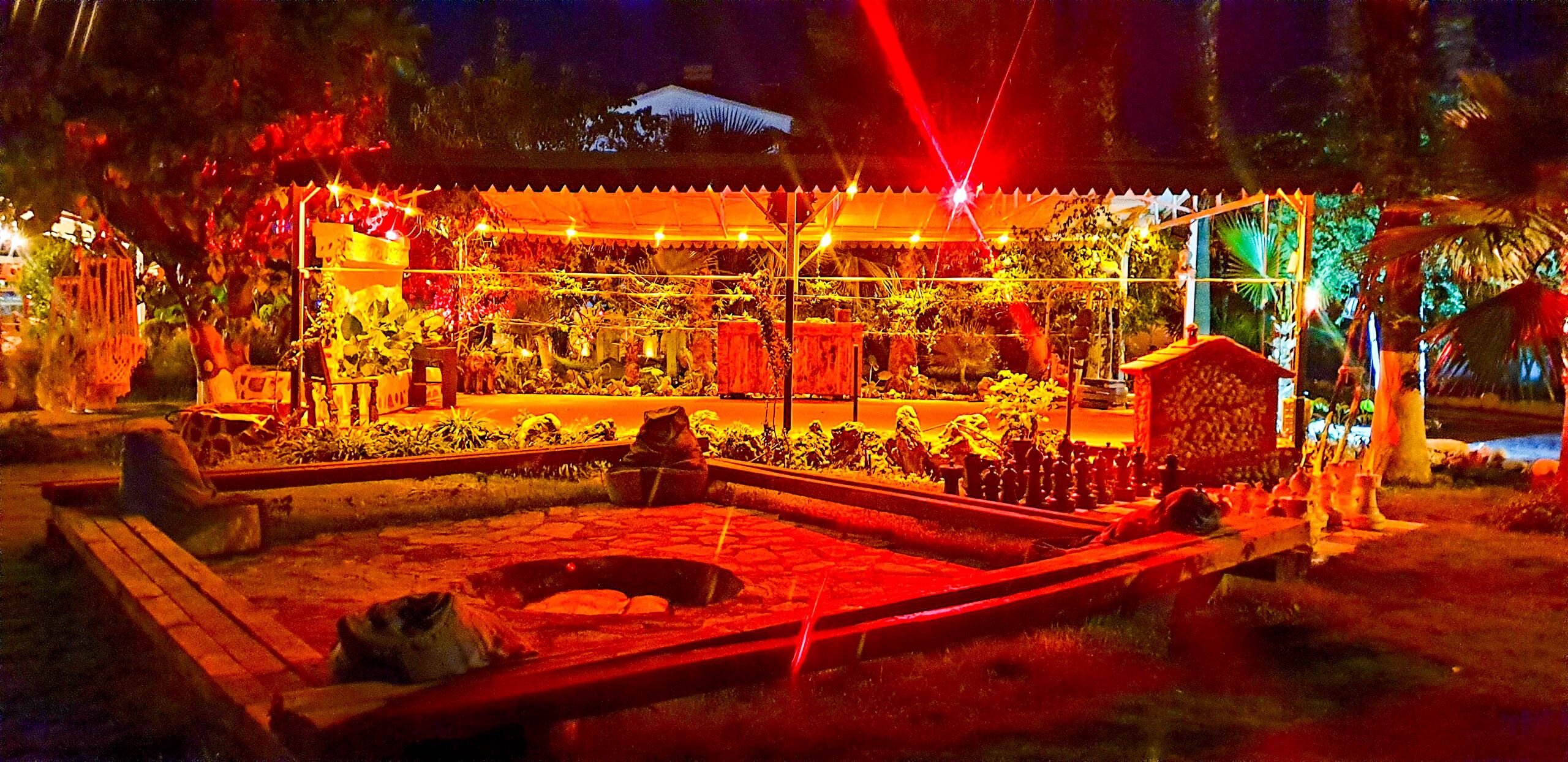 Un espace extérieur aménagé pour la nuit, doté d’une zone couverte, peut-être une terrasse ou un salon, et entouré de plantes et d’éléments décoratifs.