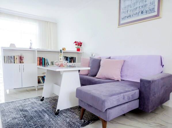 Ein modernes Wohnzimmer mit einem violetten Sofa, einem weißen Schreibtisch mit Büchern und einem Bücherregal.