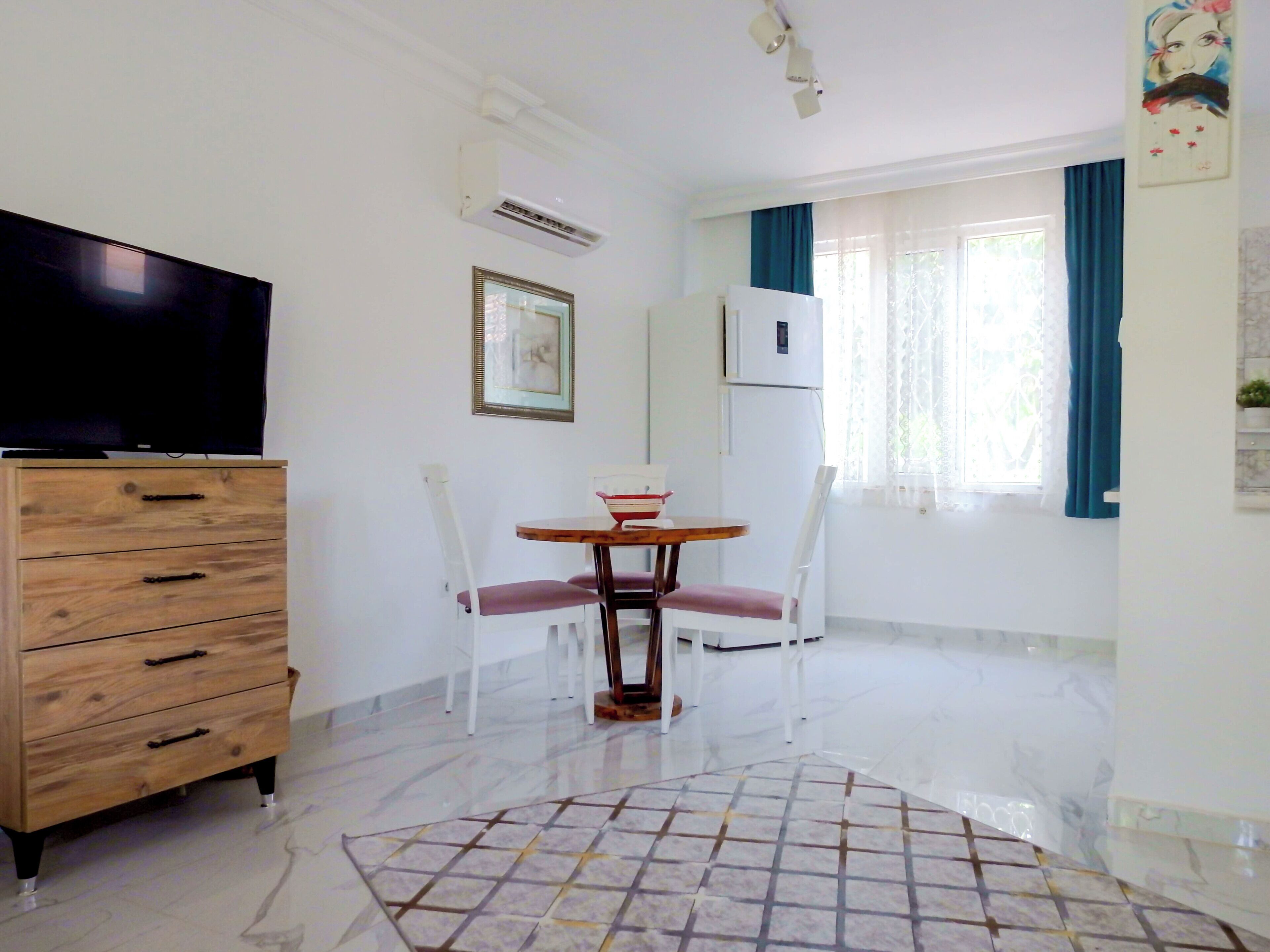 Una habitación pequeña y luminosa con un aparador de madera, un televisor, una mesa de comedor con sillas y un refrigerador.