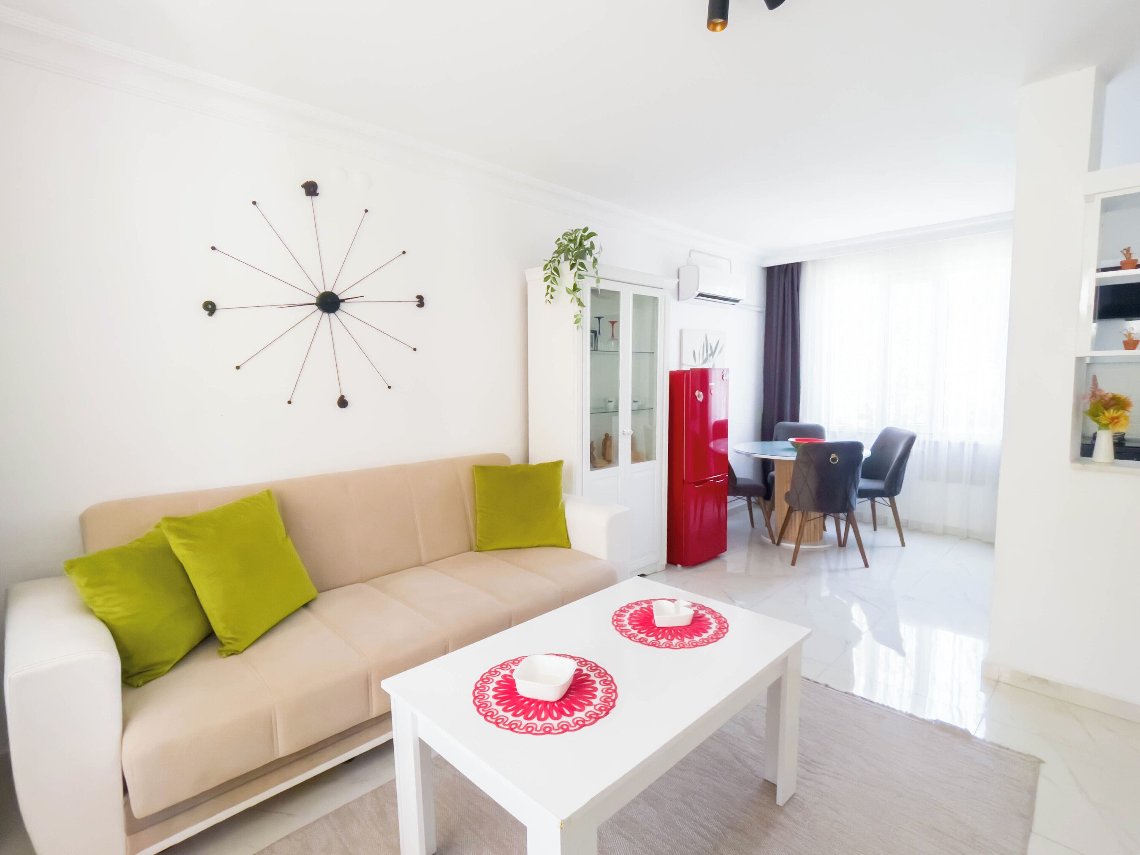 Una sala moderna con un sofá color beige, cojines verdes, una mesa de centro blanca y un refrigerador rojo, además de un comedor con mesa y sillas.