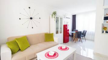 Una sala moderna con un sofá color beige, cojines verdes, una mesa de centro blanca y un refrigerador rojo, además de un comedor con mesa y sillas.