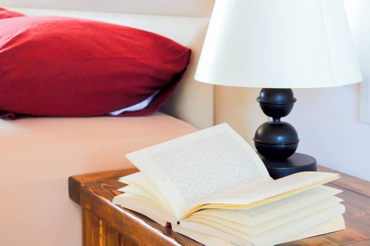 A bed with a red pillow, a bedside table with a lamp, and an open book.