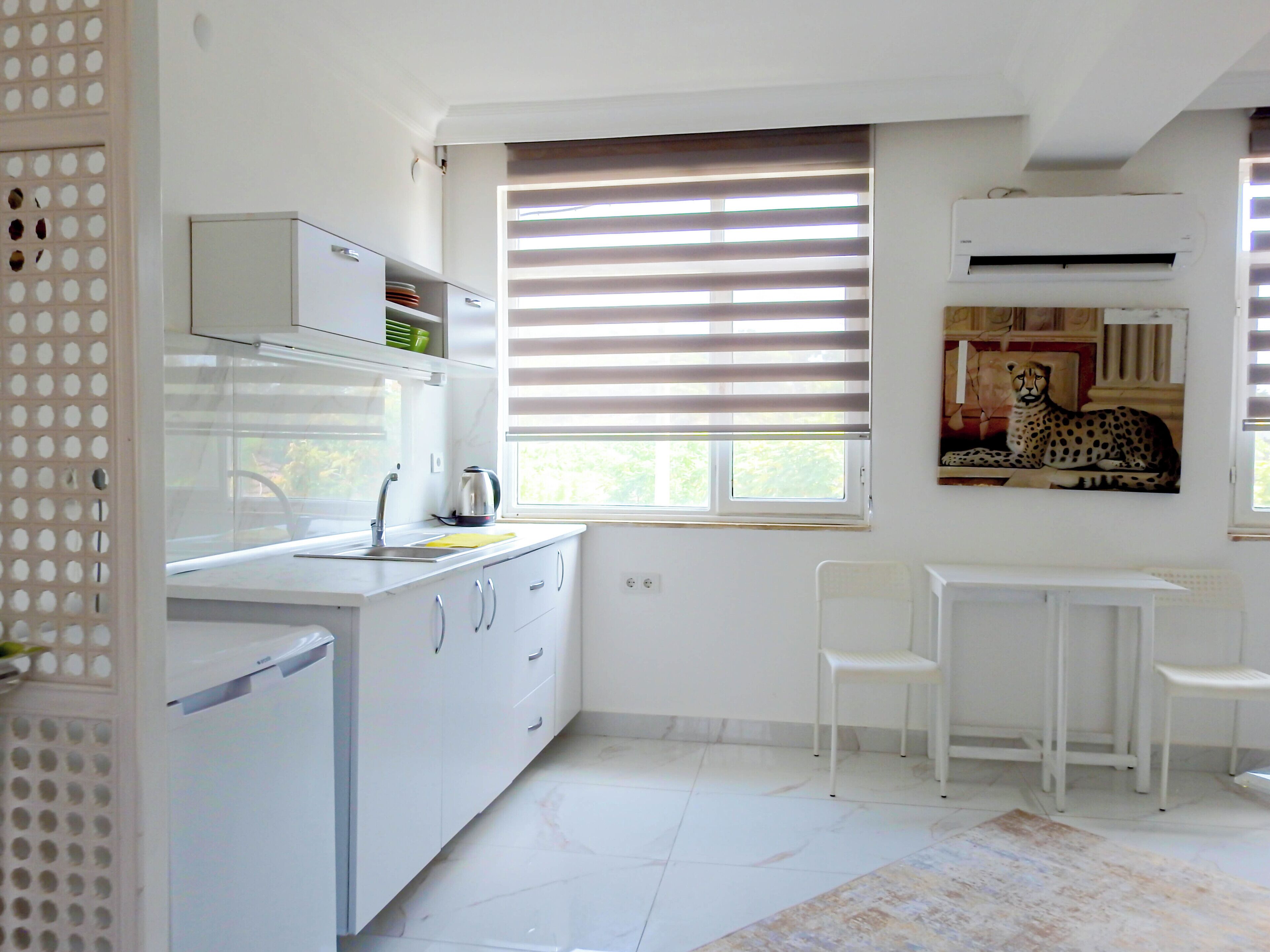 Una cocina moderna con armarios blancos, un lavamanos y un hervidor. Hay dos sillas blancas y una mesita.