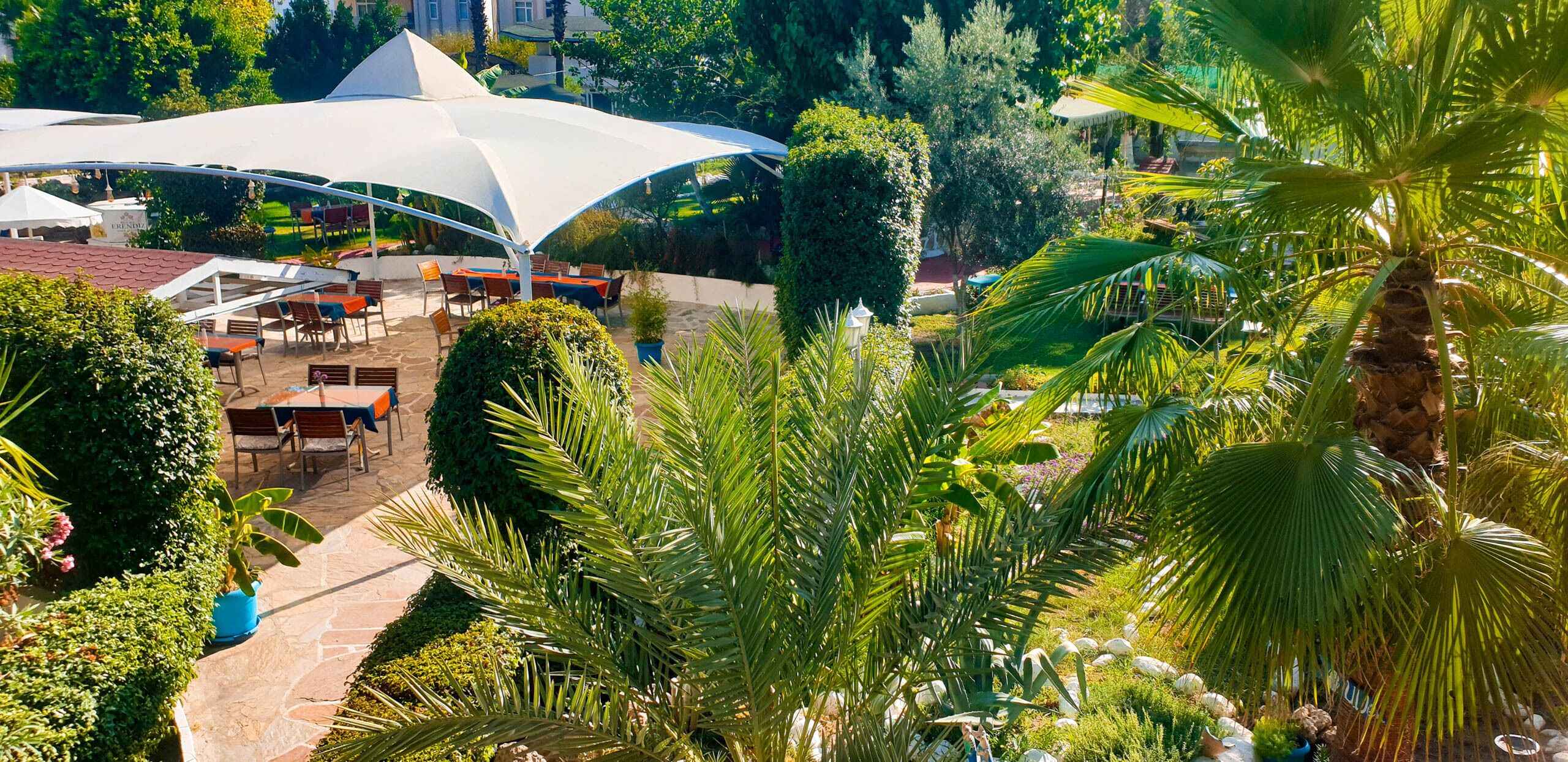 An outdoor seating area with tables and chairs under a large white canopy, surrounded by lush greenery and palm trees.
