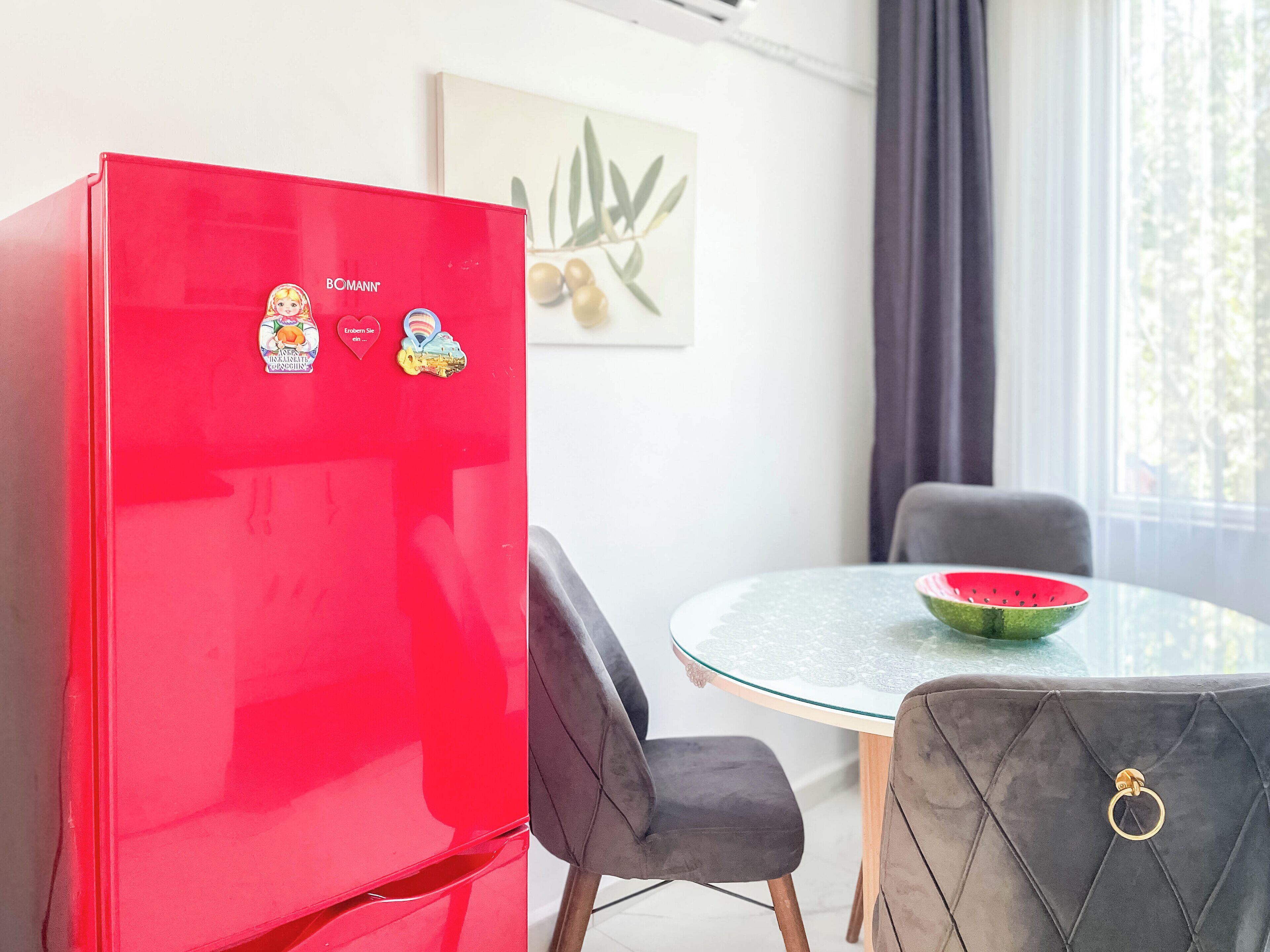 Un refrigerador rojo con calcomanías, una mesa de vidrio redonda con un cuenco de sandía y dos sillas.