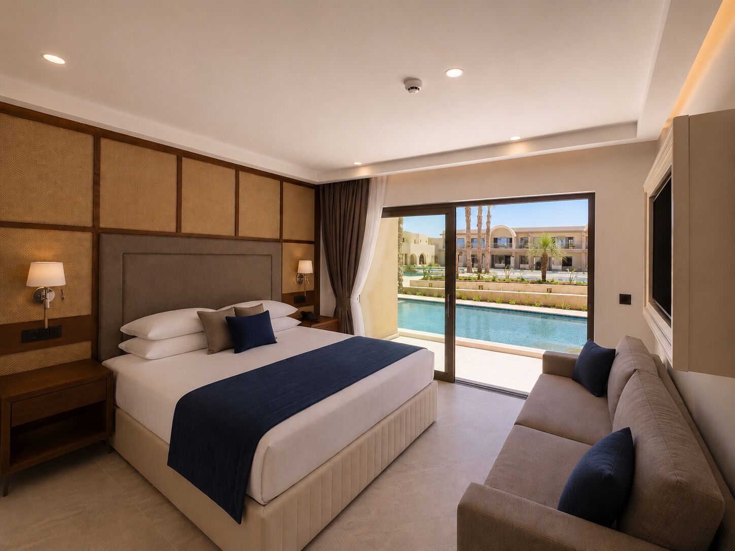 A hotel room with a bed, a nightstand, a sofa, a large window with a pool view, and a ceiling with recessed lighting.