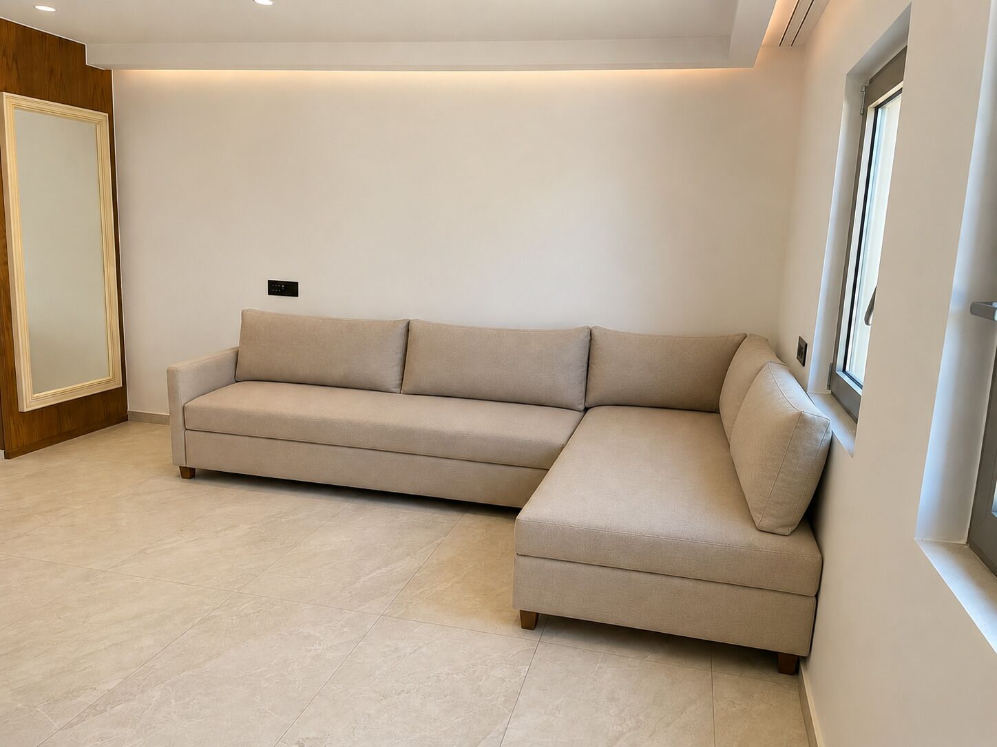 A modern living room with a beige sectional sofa, a wooden wardrobe, and a window with blinds.