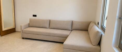 A modern living room with a beige sectional sofa, a wooden wardrobe, and a window with blinds.
