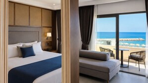 A hotel room with a bed, a sofa, a chair, a large mirror, and a view of the beach. - Nana Royal Premium Resort & Spa (Hersonissos)