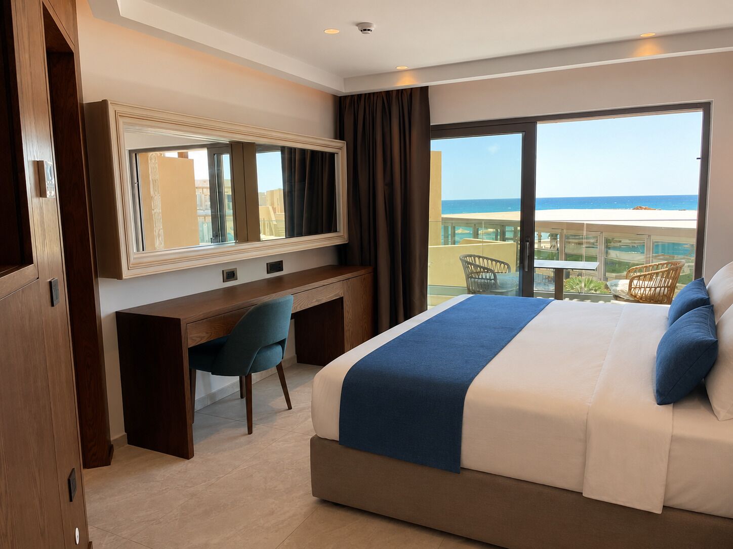 A hotel room with a bed, a desk, a chair, a large window with a view of the ocean, and a balcony with outdoor furniture.