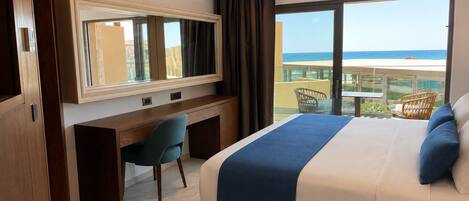 Habitación de hotel con cama, escritorio, silla, ventana grande con vista al océano y balcón con mobiliario exterior.