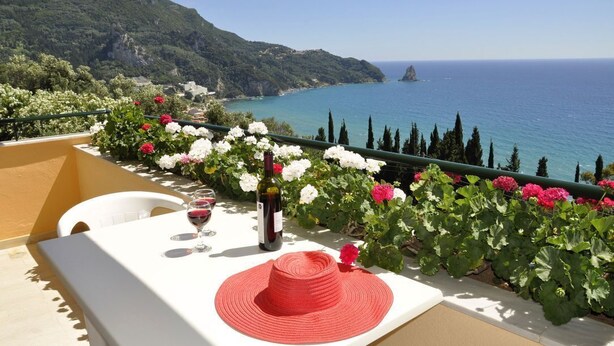 A terrace with a table set for two, a bottle of wine, and a red hat. The terrace overlooks a coastal view with flowers and cypress trees. - Pelagos Apartments (Corfu)