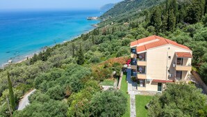A two-story building with a red-tiled roof, surrounded by lush greenery and a clear view of the sea. - Pelagos Apartments (Corfu)
