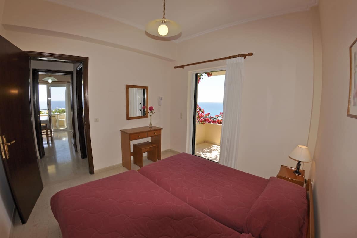 Un dormitorio con una cama grande, una mesita de noche de madera con una lámpara, un escritorio con un espejo y vistas al mar a través de una puerta de balcón.