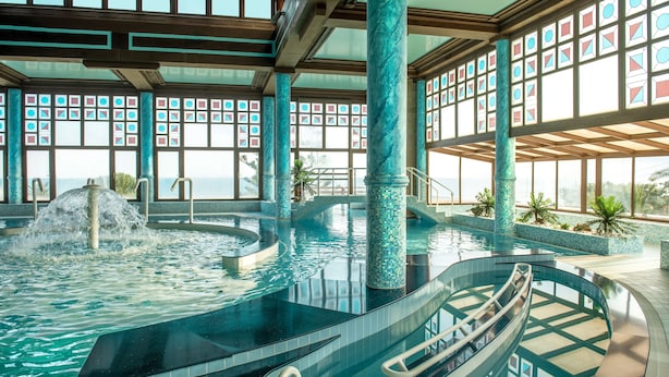 An indoor swimming pool with a waterfall feature, surrounded by large windows with geometric patterns, and a view of the ocean. - Canvas by Mitsis Cretan Village (Hersonissos)