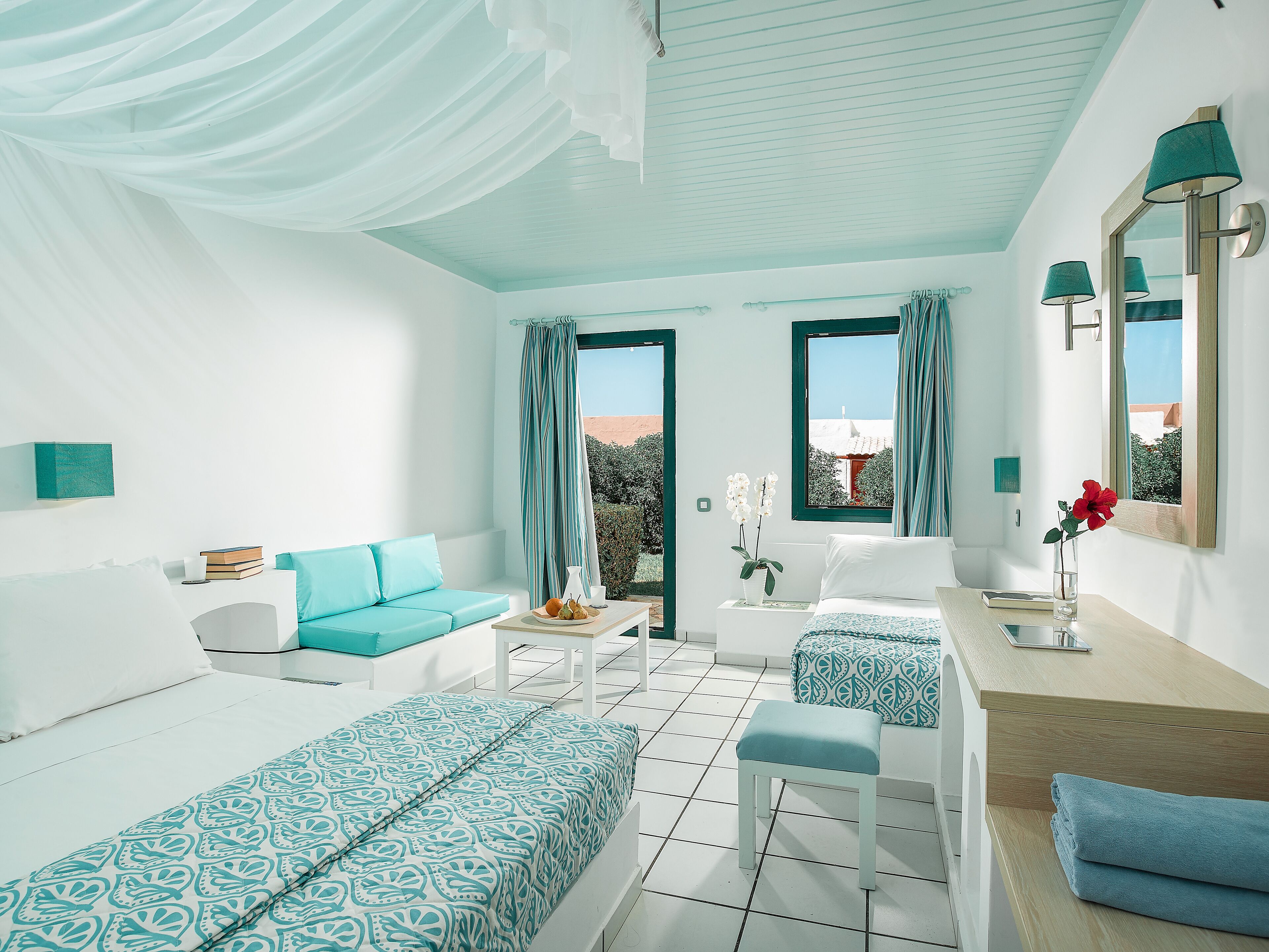 A modern hotel room with two beds, a sofa, a desk, and a view of the outdoors.