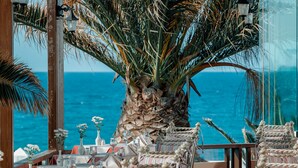 A seaside restaurant with outdoor seating, palm trees, and a view of the ocean. - Canvas by Mitsis Cretan Village (Hersonissos)