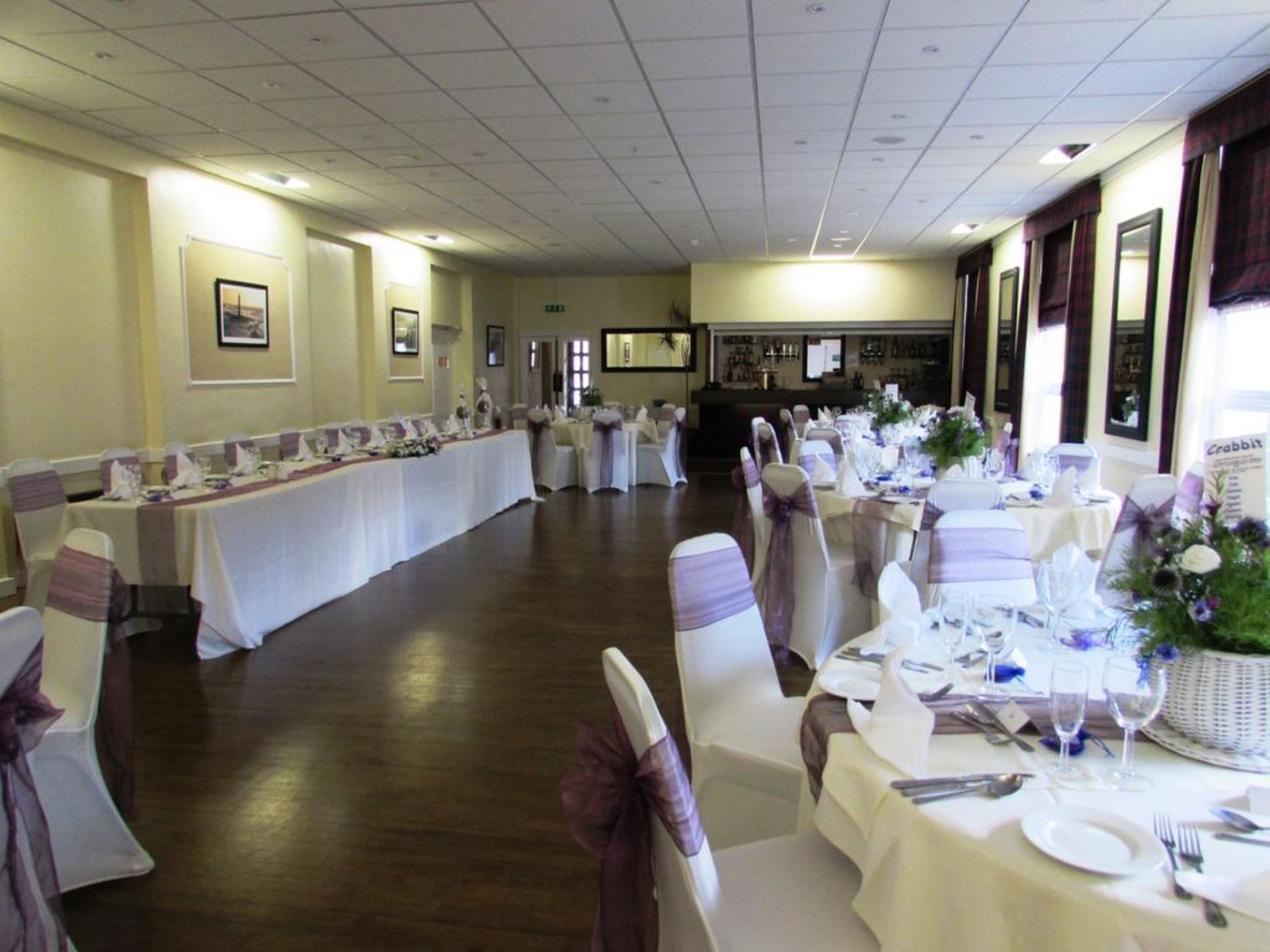 A banquet hall with long tables set for a formal event, featuring white tablecloths and purple sashes on chairs.