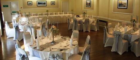 A banquet hall with round tables, white chairs with grey sashes, and framed pictures on the walls.