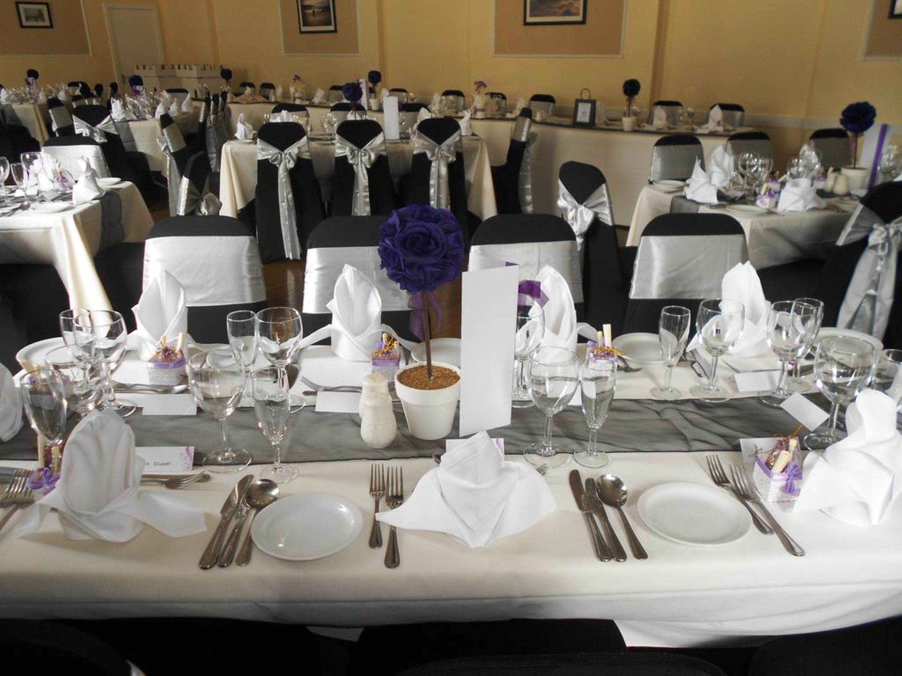 Un salón de banquetes con mesas dispuestas para un evento formal, con manteles blancos, sillas negras adornadas con cintas plateadas, y copas y cubiertos ordenados.
