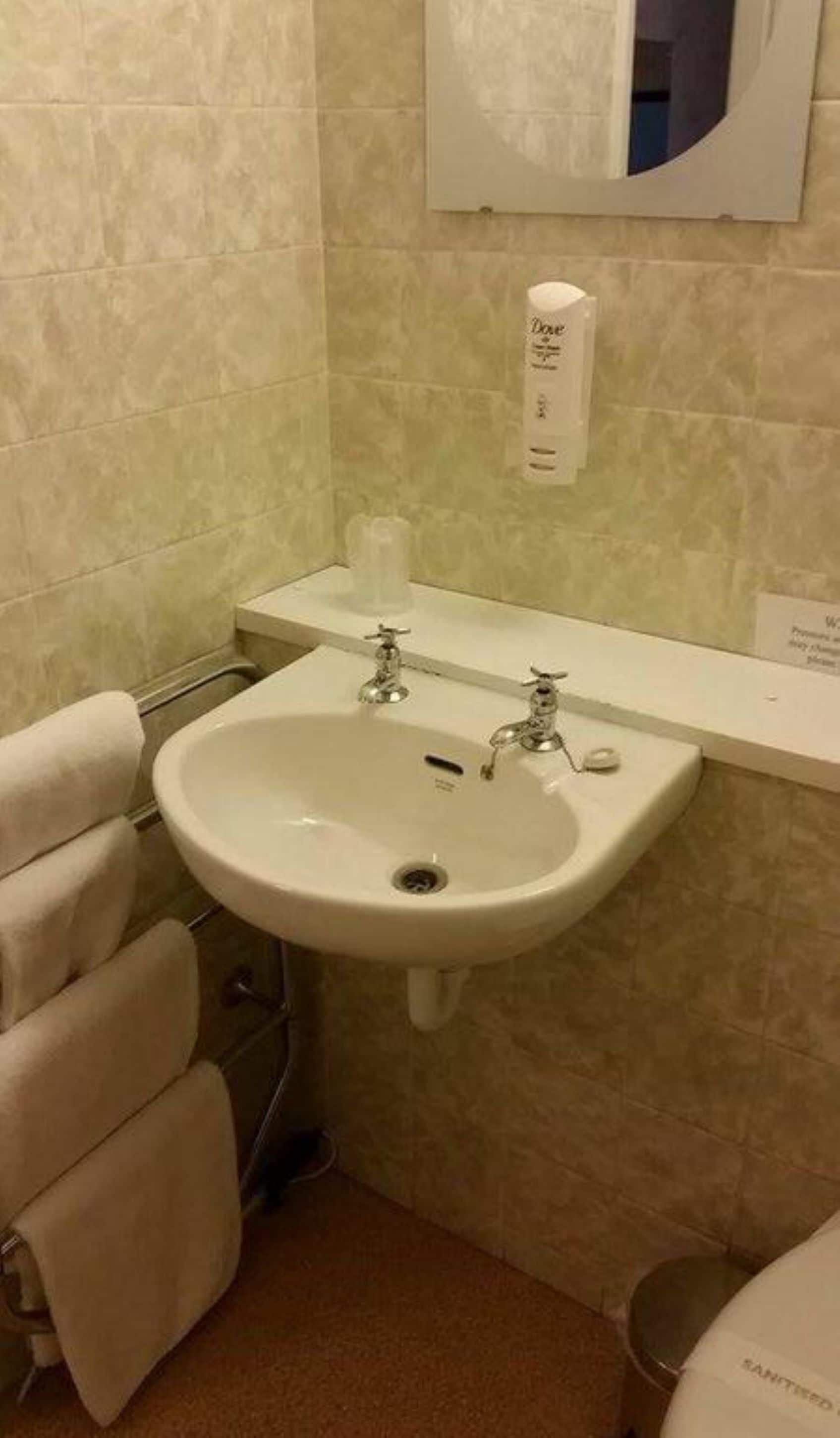 A bathroom with a white sink, two faucets, a towel rack, and a hand sanitizer dispenser on the wall.