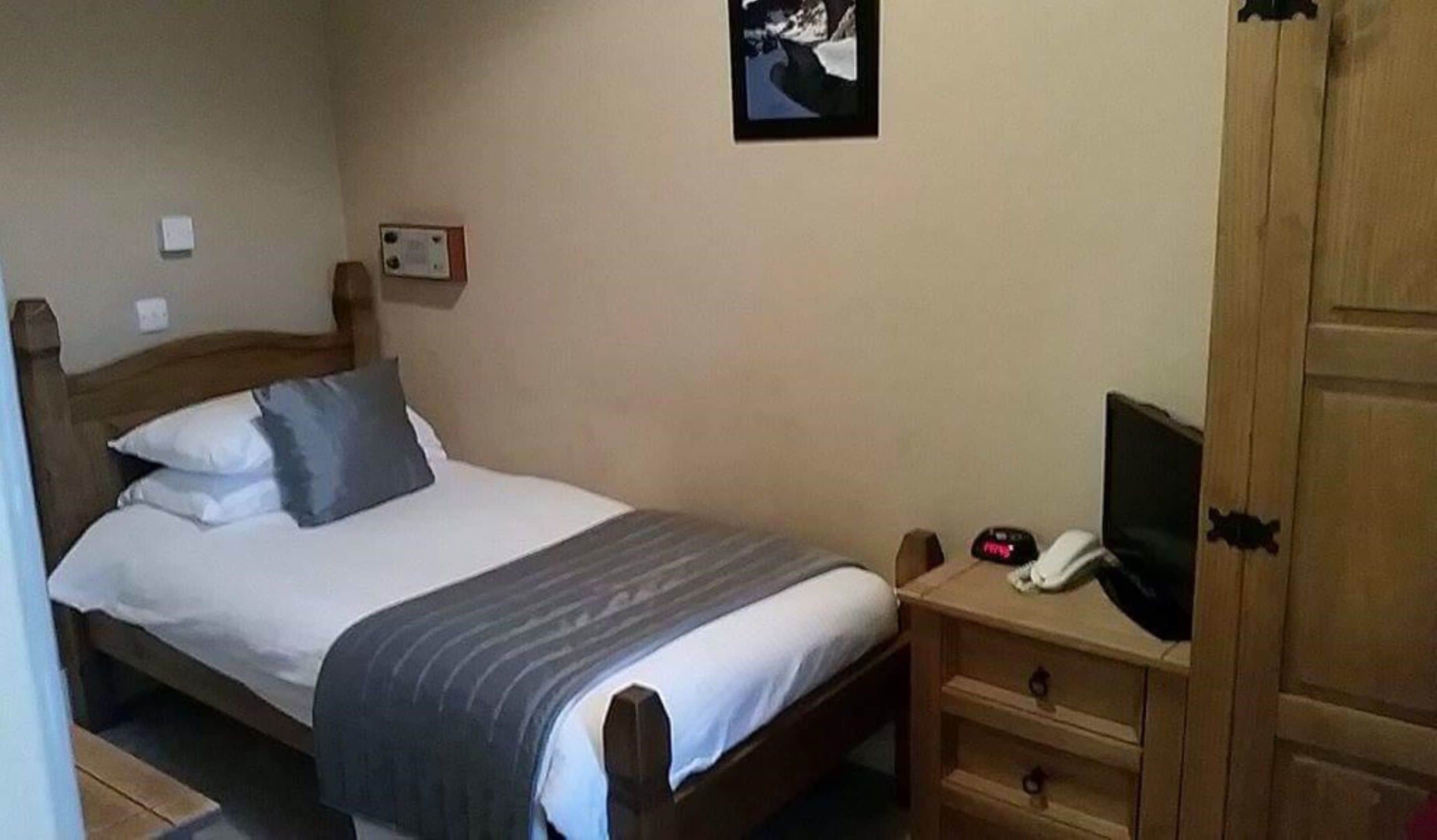 Uma cama de solteiro com um cobertor cinzento, uma mesa de cabeceira com um telefone e um pequeno alto-falante, e um guarda-roupa de madeira.