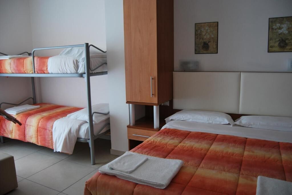 A dormitory room with bunk beds, a wooden wardrobe, and a single bed with a colorful blanket.