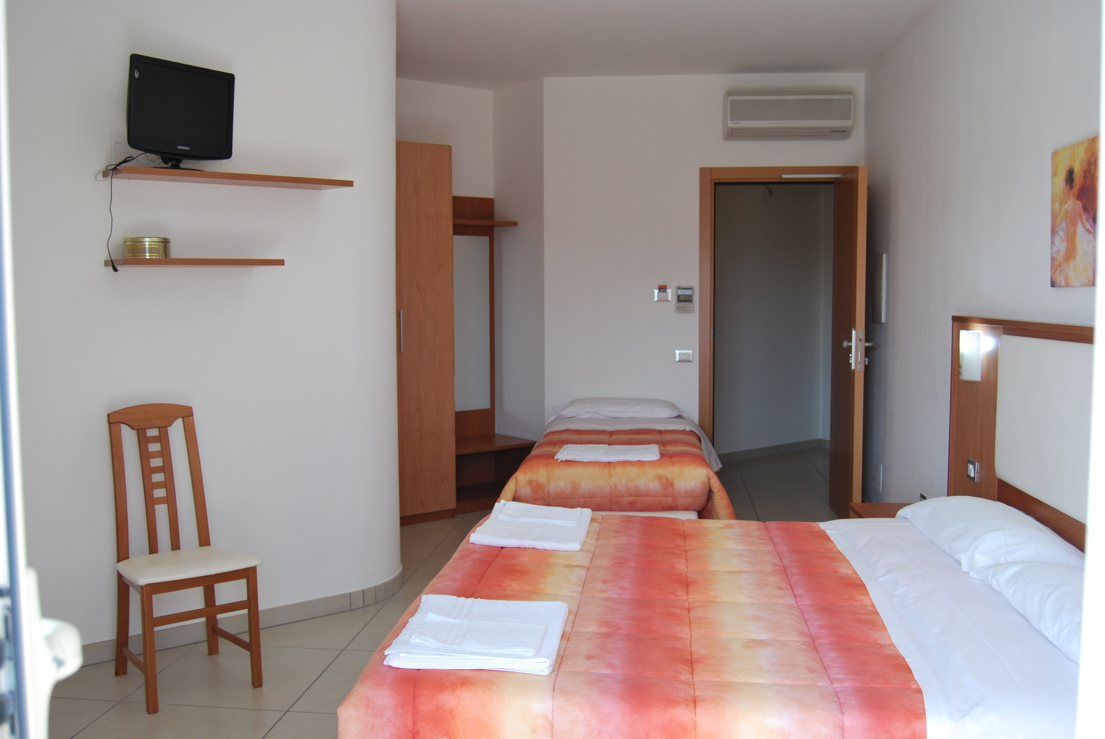 Quarto de hotel com duas camas, uma televisão em uma prateleira, uma cadeira e uma porta.