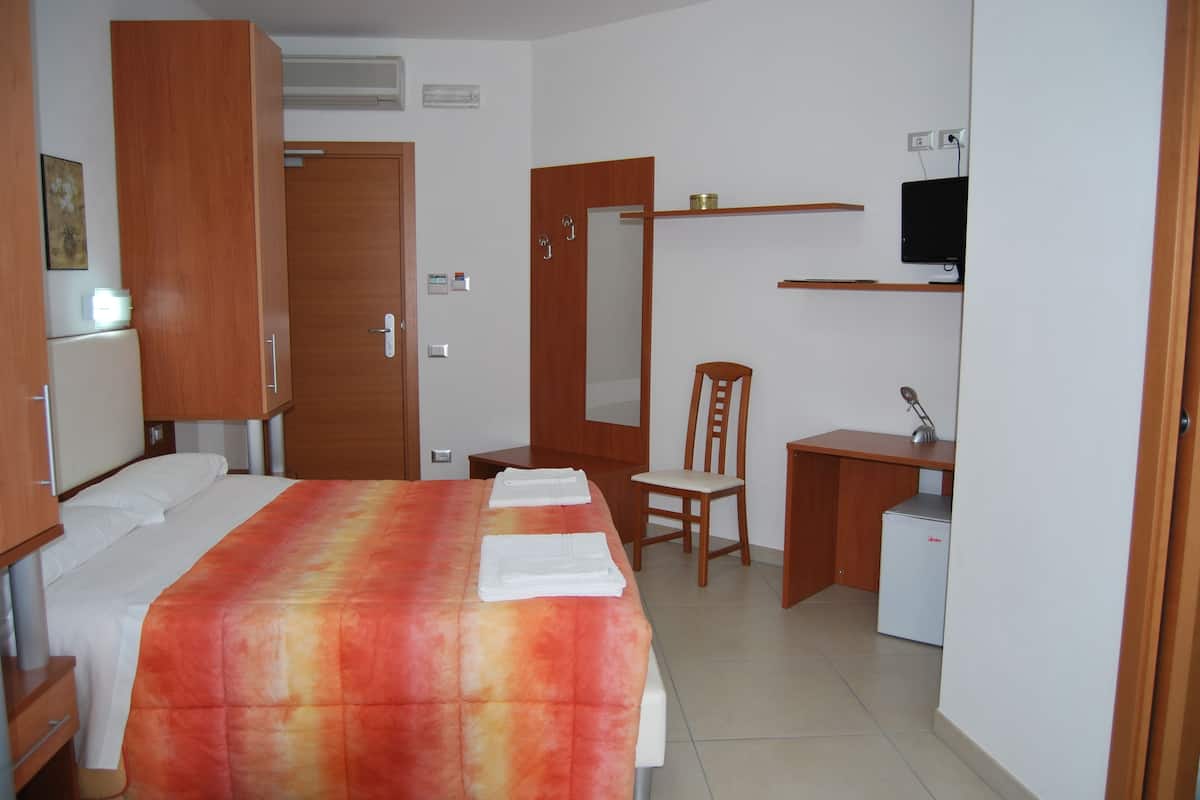 Quarto de hotel com cama, escrivaninha, cadeira, televisão e um pequeno frigorífico.