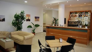 Un vestíbulo de hotel con mostrador de recepción, zona de bar, área de estar con sofá y sillones, y plantas en macetas.