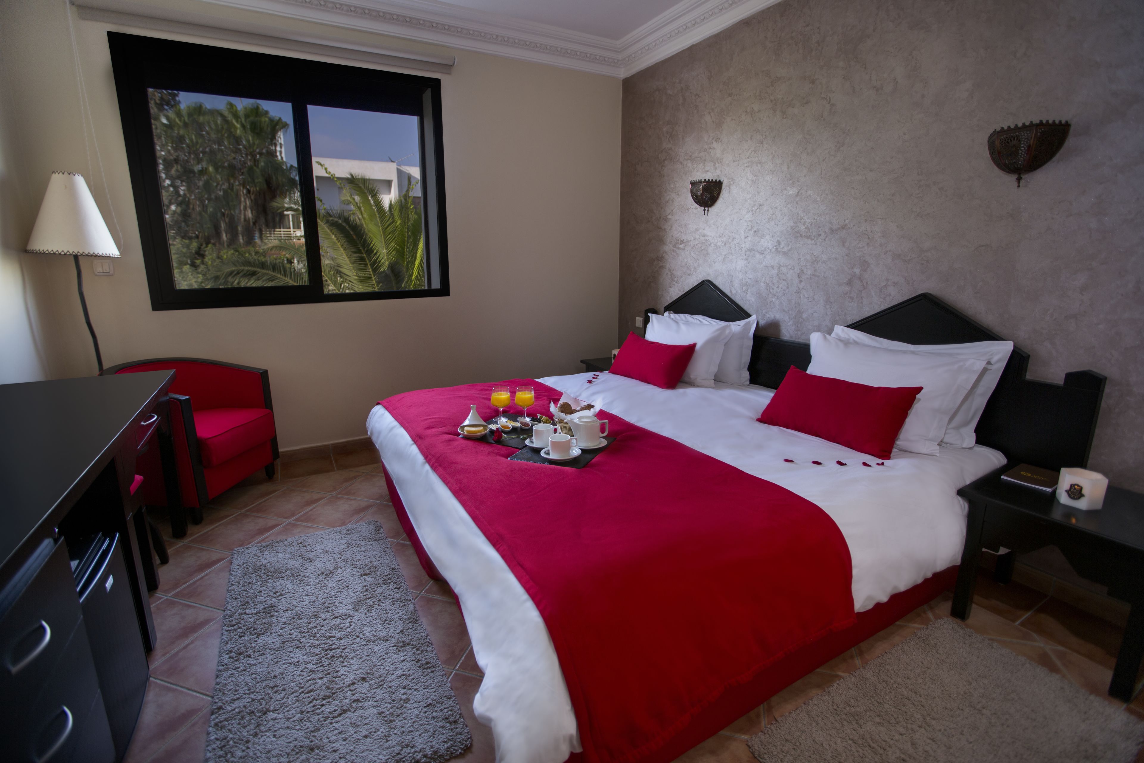 Habitación de hotel con una cama grande, un escritorio, una silla, una mesita de noche y un ventanal con vista a palmeras.