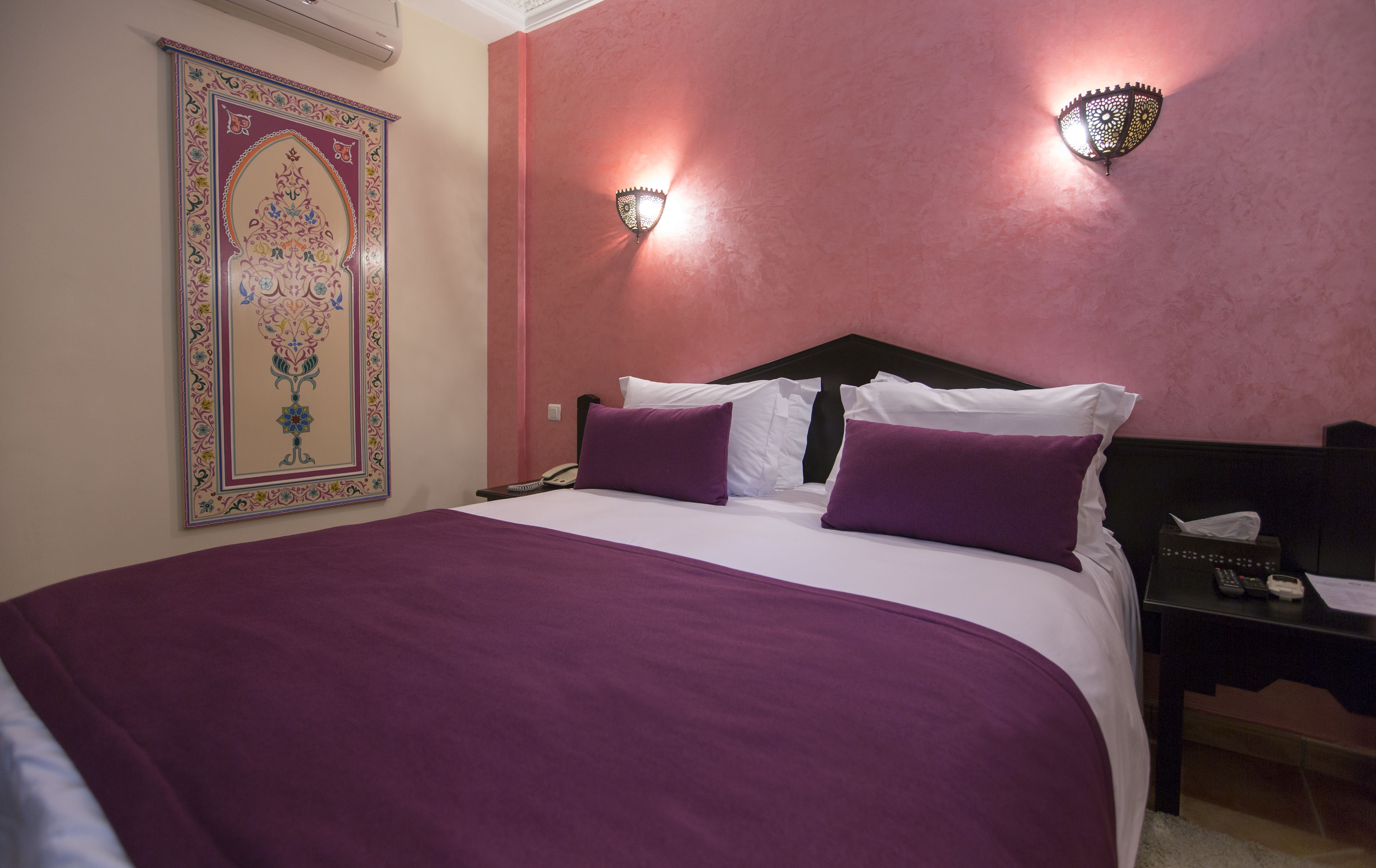 A hotel room with a large bed, purple bedding, a decorative wall hanging, and two wall-mounted light fixtures.