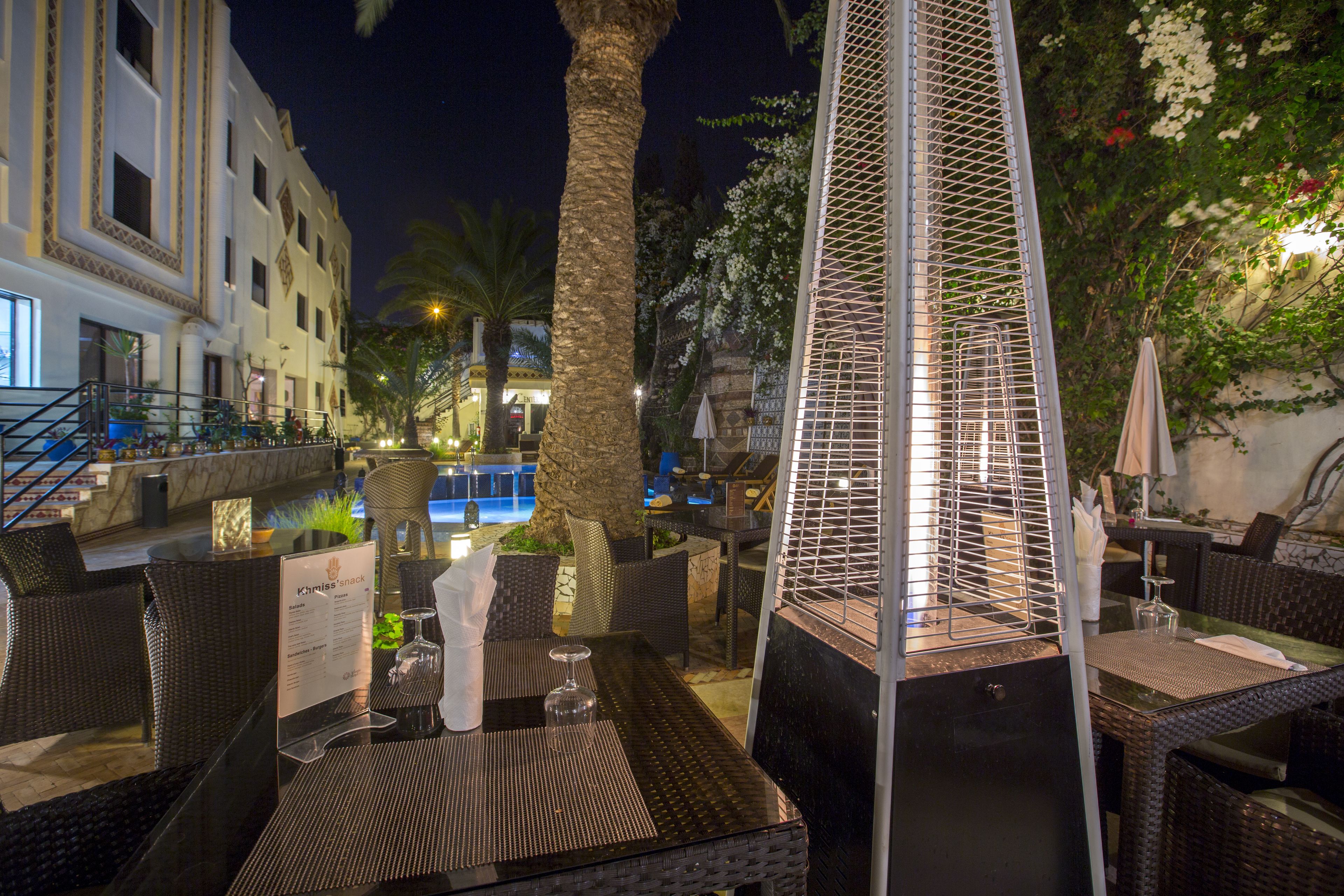 Un área de comedor al aire libre con mobiliario de mimbre, una piscina al fondo y una estructura metálica decorativa.