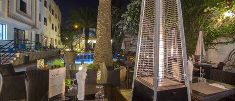 Un área de comedor al aire libre con mobiliario de mimbre, una piscina al fondo y una estructura metálica decorativa.