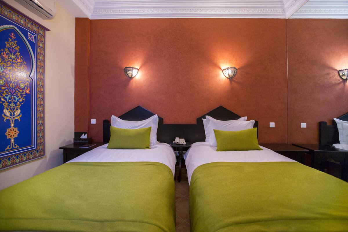 A hotel room with two beds, each with green bedding and white pillows. There are wall sconces with decorative lamps, a telephone on the nightstand, and a decorative wall hanging.