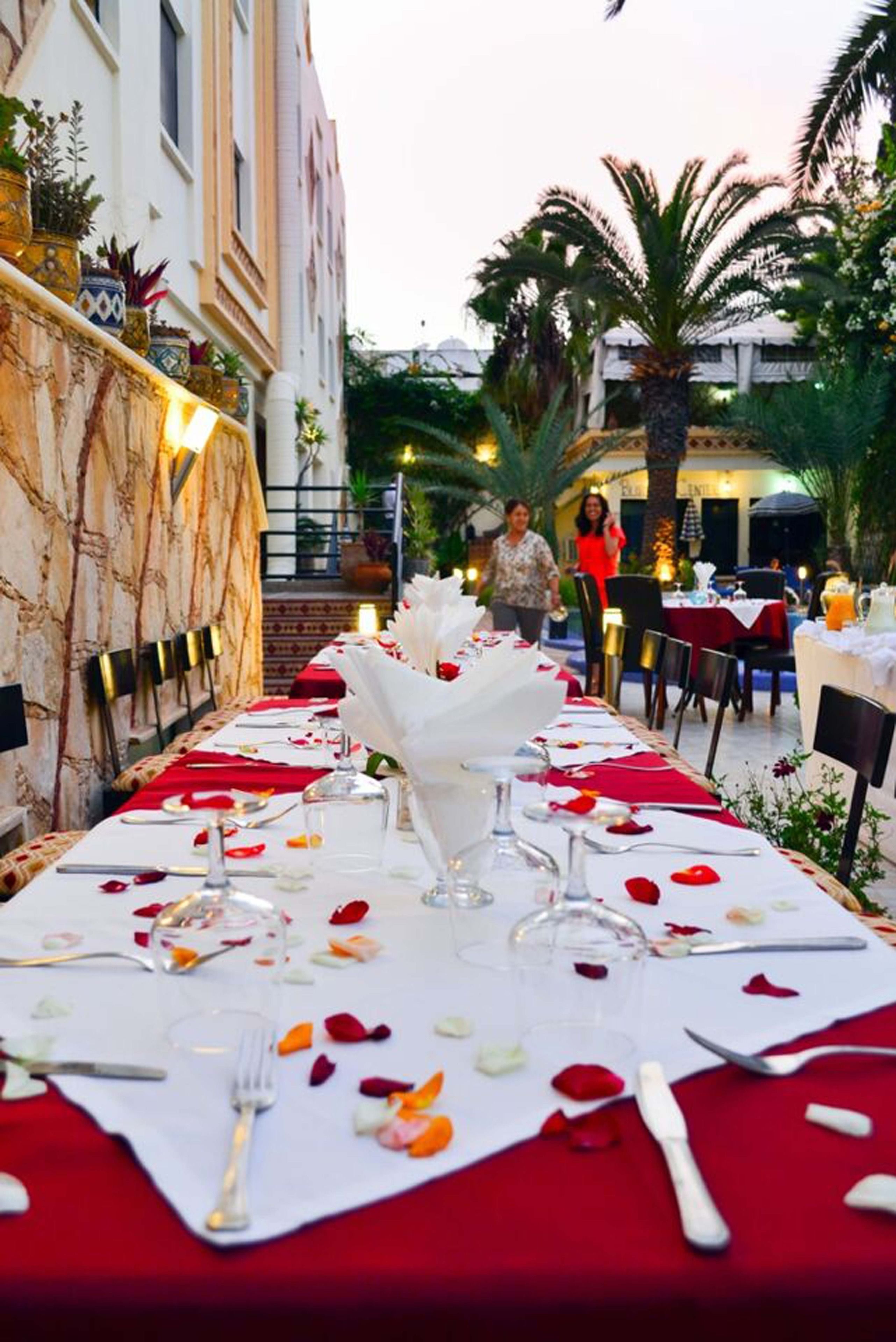 An outdoor dining area with a long table set for a meal, surrounded by buildings and palm trees.
