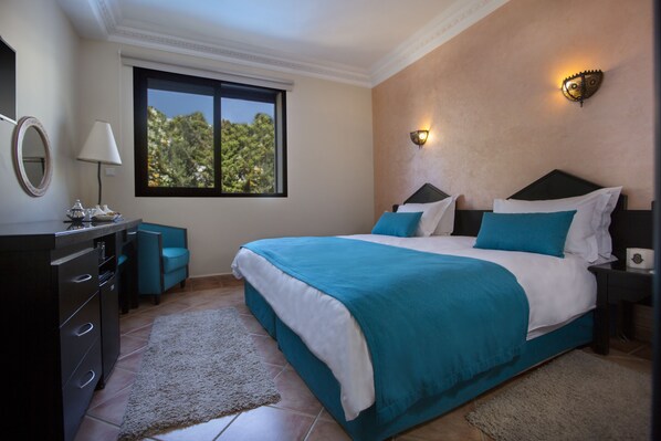 A bedroom with a large bed, a dresser, a nightstand, a lamp, and a window with a view of trees. - Atlantic Hotel Agadir (Agadir)