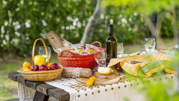 Ein Picknick mit einer Korb voller Früchte, einer Weinflasche und Käse auf einem Holztisch im Freien.