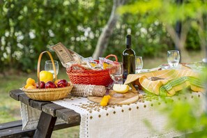 A picnic setup with a basket of fruits, a bottle of wine, and a cheese on a wooden table outdoors.