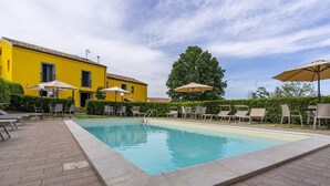 Ein Pool mit Liegestühlen und Sonnenschirmen, umgeben von einem gelben Gebäude und Grünflächen.