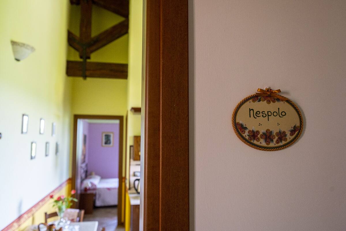 A room with a wooden ceiling, a bed, a table with chairs, and a wall-mounted sign that reads "nespolo."